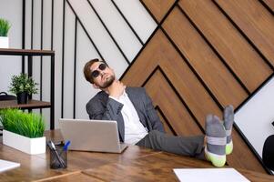 une homme est séance dans le Bureau avec une ordinateur portable, le sien pieds sont sur le tableau. tergiverser photo