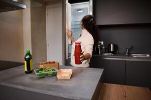 Jeune femme tirant en dehors une verre bouteille avec rouge jus de tomates de le réfrigérateur. femme au foyer en train de préparer en bonne santé dîner. papier carton recyclable des boites avec Frais légumes sur le cuisine compteur photo