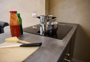 encore la vie inoxydable acier casserole sur le surface de électrique le fourneau et Frais Ingrédients sur le cuisine tableau. bouteille avec tomate jus ou passata, une portion de fromage sur une Coupe planche photo