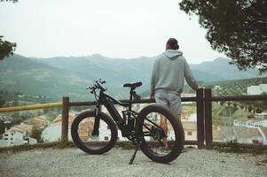 arrière vue de une Jeune actif homme cycliste permanent près le sien électrique batterie alimenté vélo, admiratif magnifique montagnes, profiter équitation dans le la nature photo