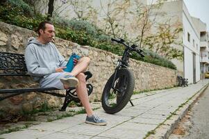 Jeune adulte cycliste relaxant lorsque équitation électrique bicyclette dans le ville, en portant une bouteille de l'eau tandis que séance sur le banc. actif personnes. Urbain mode de vie. respectueux de la nature transport. photo