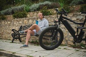 Jeune homme cycliste dans tenue de sport, en buvant l'eau de des sports bouteille dans le ville, séance sur une banc, relaxant après équitation un respectueux de la nature vélo électrique moto. électrique vélo dans le premier plan photo