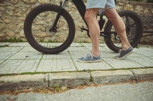 tondu vue jambes de une musclé sportif homme pousser le sien électrique Montagne bicyclette dans le ville. aventure Voyage sur vélo. en utilisant vélo électrique comme respectueux de la nature transport photo