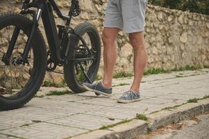 fermer jambes de une cycliste dans des sports chaussures, pousser le sien bicyclette tandis que en marchant le long de le rue. homme en utilisant vélo électrique comme écologique durable mode de Urbain économique transport. ville vie. personnes. mode de vie. photo