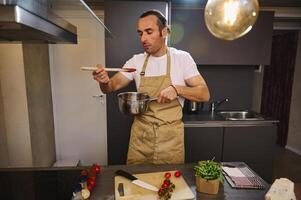 Beau caucasien homme dégustation tomate sauce, permanent à le fourneau dans le moderne intérieur de Accueil cuisine. Masculin chef cuisine passata selon à traditionnel italien recette photo