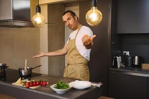 Beau caucasien homme dans du chef tablier cuisine famille dîner, souriant et montrant le cuisine compteur avec Frais Ingrédients à la recherche à caméra photo