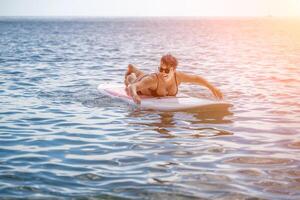 femme souper mer Voyage. des sports fille sur une planche de surf dans le mer sur une ensoleillé été journée. dans une noir baignade costume, il est assis sur une sapa dans le mer. du repos sur le mer. photo