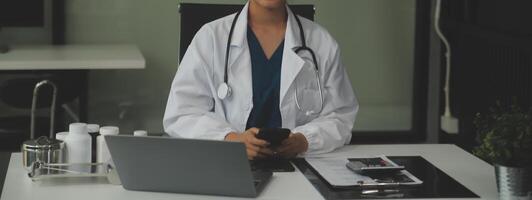 sérieux femelle médecin en utilisant portable et l'écriture Remarques dans médical journal séance à bureau. Jeune femme professionnel médical médecin portant blanc manteau et stéthoscope travail sur ordinateur à lieu de travail. photo