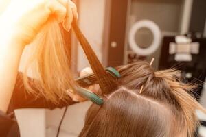le coiffeur prépare une femme, fait du une coiffure avec boucles en utilisant une curling fer. longue lumière marron Naturel cheveux. soi se soucier concept. photo