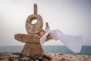 une femme est assis sur une pierre sculpture fabriqué de grand des pierres. elle est habillé dans une blanc longue robe, contre le toile de fond de le mer et ciel. le robe développe dans le vent. photo