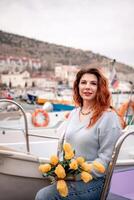 femme détient Jaune tulipes dans port avec bateaux amarré dans le arrière-plan., couvert jour, Jaune chandail, montagnes photo