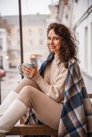 une âge moyen femme dans une beige chandail avec une bleu agresser dans sa mains est dans une rue café sur le véranda photo