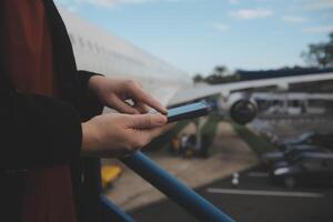 Jeune asiatique femme dans international aéroport, en utilisant mobile téléphone intelligent et vérification vol à le vol information planche photo
