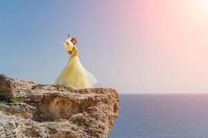 femme dans une Jaune robe sur le mer. côté vue Jeune magnifique sensuel femme dans Jaune longue robe posant sur une Roche haute au dessus le mer à le coucher du soleil. fille dans la nature contre le bleu ciel photo