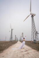 une femme dans une blanc robe est en marchant vers le bas une saleté route dans de face de une rangée de vent turbines. photo