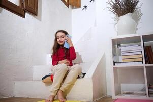 magnifique élémentaire âge école fille ayant une conversation sur Téléphone avec sa amis. peu enfant fille parlant sur mobile téléphone, séance sur pas dans le confortable rural maison intérieur. photo