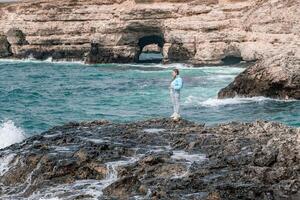 une femme dans une bleu veste des stands sur une Roche au dessus une falaise au dessus le mer et regards à le rage océan. fille voyageur repose, pense, rêves, jouit la nature. paix et calme paysage, venteux temps. photo