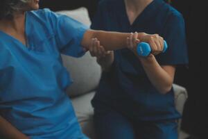 vieux femme formation avec physiothérapeute en utilisant haltères à maison. thérapeute assistant Sénior femme avec des exercices dans allaitement maison. personnes âgées patient en utilisant haltères avec tendu bras. photo