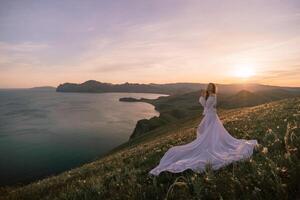 une femme dans une blanc robe des stands sur une colline surplombant une corps de l'eau. le scène est serein et pacifique, avec le Soleil réglage dans le Contexte. le femme est perdu dans pensée. photo