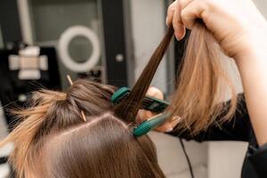 le coiffeur prépare une femme, fait du une coiffure avec boucles en utilisant une curling fer. longue lumière marron Naturel cheveux. soi se soucier concept. photo
