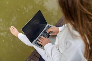 une femme est séance sur une Dock avec une portable ouvert dans de face de son. elle est dactylographie sur le clavier et elle est concentré sur sa travail. concept de productivité et concentration. photo