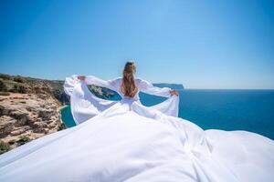 blond avec longue cheveux sur une ensoleillé rivage dans une blanc écoulement robe, arrière voir, soie en tissu agitant dans le vent. contre le toile de fond de le bleu ciel et montagnes sur le rivage. photo