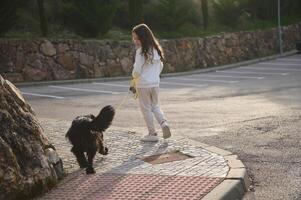 plein longueur portrait de une magnifique peu enfant fille dans tenue de sport, en marchant sa chien sur laisse sur le rue en plein air. gens et animaux. en jouant animaux domestiques concept photo