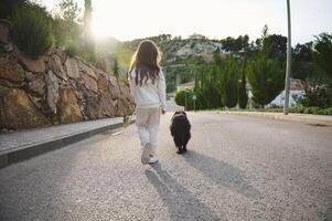 vue de le retour de une mignonne enfant fille dans blanc des sports vêtements en marchant sa chien sur laisse sur le campagne contre le coucher du soleil rayons de soleil Contexte. personnes, en jouant animaux domestiques et la nature concept photo