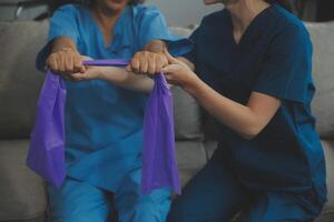 personnel entraîneur assistant Sénior femme avec la résistance groupe. réhabilitation physiothérapie ouvrier portion vieux patient à allaitement maison. vieux femme avec étendue bande étant coaché par physiothérapeute. photo