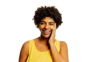 de bonne humeur magnifique Jeune femme avec frisé cheveux et un appareil dentaire sur les dents en riant et émouvant sa visage par main isolé plus de blanc Contexte photo