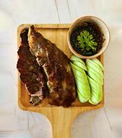 Haut vue de grillé porc côtes, tranché concombre et épicé sauce sur en bois plateau. plat allonger de barbecue nourriture avec Frais des légumes sur blanc marbre. photo