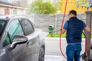 Jeune homme lavages le sien voiture à une en libre service voiture laver en utilisant une tuyau avec pressurisé l'eau et mousse. photo