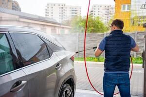 Jeune homme lavages le sien voiture à une en libre service voiture laver en utilisant une tuyau avec pressurisé l'eau et mousse. photo