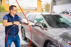 Jeune homme lavages le sien voiture à une en libre service voiture laver en utilisant une tuyau avec pressurisé l'eau et mousse. photo