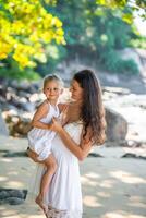 Jeune femme mère avec une peu fille dans blanc Robes sur rivage dans le ombre de des arbres et paumes photo