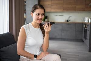 Jeune femme parle une voix message sur sa téléphone intelligent tandis que séance sur le canapé à Accueil photo