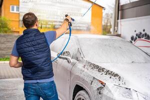 Jeune homme lavages le sien voiture à une en libre service voiture laver en utilisant une tuyau avec pressurisé l'eau et mousse. haute qualité photo