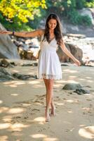 élégant attrayant Jeune femme portant dans blanc robe ayant amusement sur sur rivage dans le ombre de des arbres et palmiers, liberté et bonheur photo