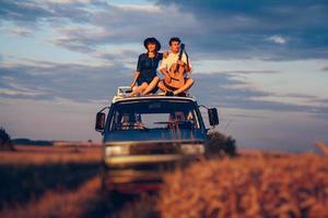 homme couple avec une guitare et une femme au chapeau sont assis sur le toit d'une voiture dans un champ de blé photo