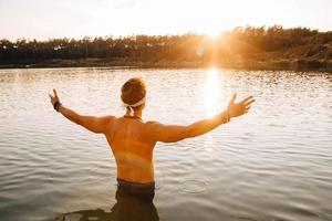homme avec une musculature parfaite se tient dans l'eau au coucher du soleil, jetant ses mains sur le côté photo