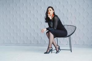 femme portant une robe noire est assise à la chaise en studio blanc photo