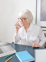 femme senior aux beaux cheveux gris en blouse blanche buvant de l'eau pendant le travail au bureau. travail, personnes âgées, bilan hydrique, trouver une solution, concept d'expérience