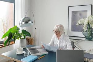 femme âgée fatiguée de beaux cheveux gris en blouse blanche lisant des documents au bureau. travail, personnes âgées, problèmes, trouver une solution, concept d'expérience