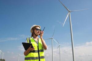 le sur de soi femme dans blanc casque travail avec numérique tablette et en utilisant walkie talkie à une renouvelable énergie cultiver. femelle inspecteur contrôler le fonctionnement de vent turbines en plein air photo