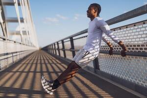 Jeune Afro-américain homme est exercice sur le pont dans le ville. il est Faire sens inverse des pompes. photo