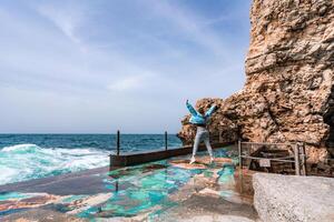 une femme dans une bleu veste des stands sur une Roche au dessus une falaise au dessus le mer et regards à le rage océan. fille voyageur repose, pense, rêves, jouit la nature. paix et calme paysage, venteux temps. photo