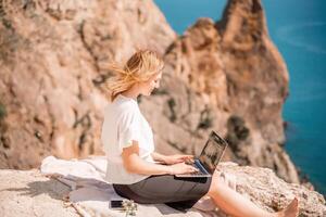 free-lance femme travail sur une portable par le mer, dactylographie une façon sur le clavier tandis que profiter le magnifique voir, mise en évidence le idée de éloigné travail. photo