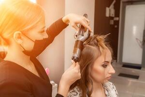 une coiffeur dans une noir masque prépare une femme, fait du une coiffure avec boucles en utilisant une curling fer. longue lumière marron Naturel cheveux. soi se soucier concept. photo