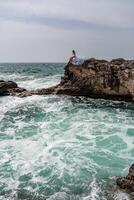 une femme dans une orage est assis sur une pierre dans le mer. habillé dans une blanc longue robe, vagues crash contre le rochers et blanc vaporisateur monte. photo