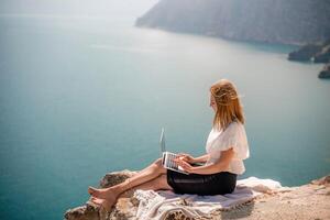 free-lance femme travail sur une portable par le mer, dactylographie une façon sur le clavier tandis que profiter le magnifique voir, mise en évidence le idée de éloigné travail. photo